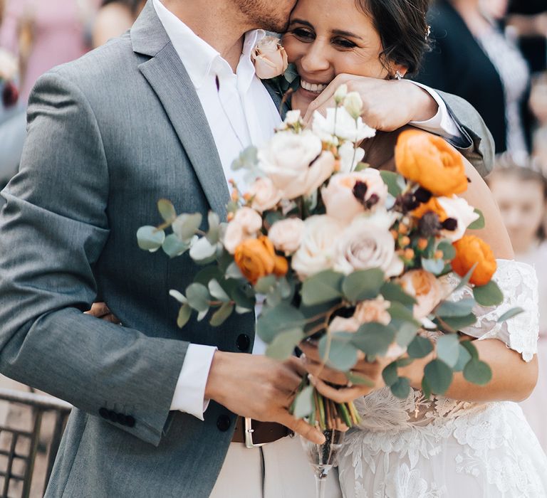 Groom in grey suit jacket kisses the bride on the cheek as she holds orange and white wedding bouquet with peonies and roses