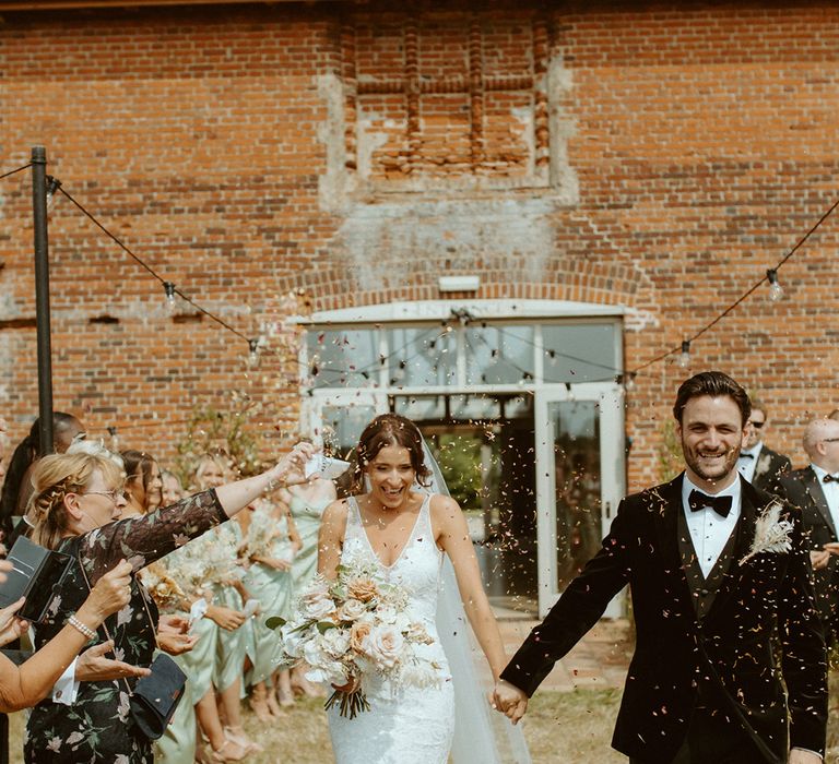 Bride in lace wedding dress with front slit holds hands with groom in black tie as the guests throw confetti