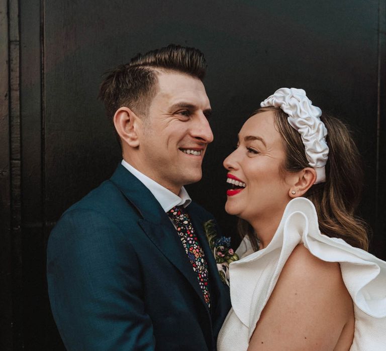 Smiling groom in bright floral tie and bride wearing red lipstick, large ruffle headband and wedding dress with statement sleeves