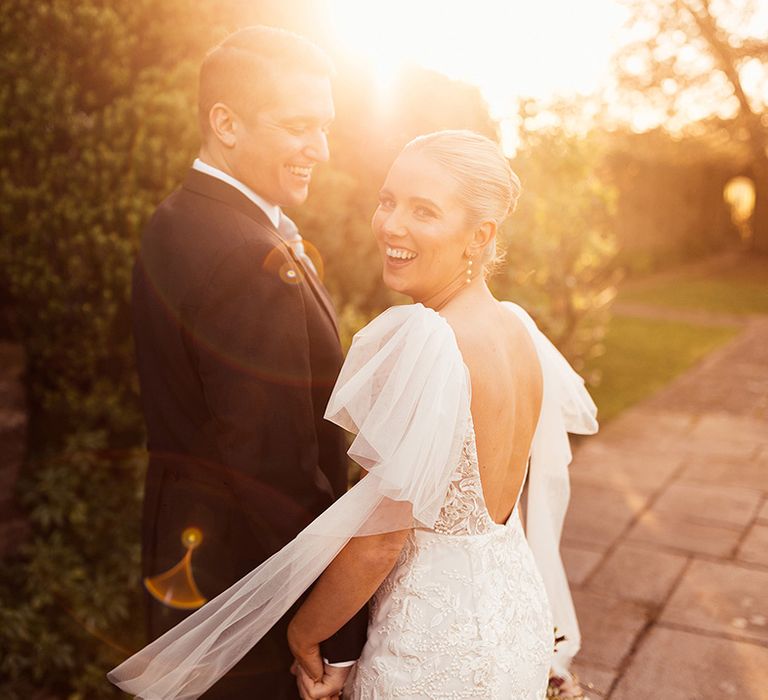 Bride in lace scoop back wedding dress with ruffle sleeves and pearl drop earrings holds hands with groom during golden hour 