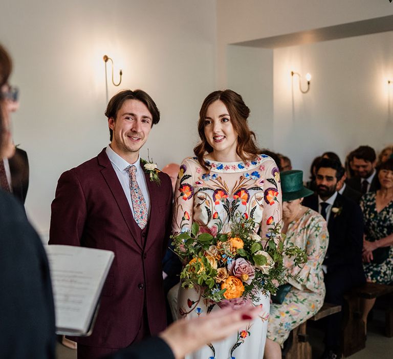 Groom in three piece burgundy suit with blue suit and floral tie with bride in bright and colourful floral wedding dress