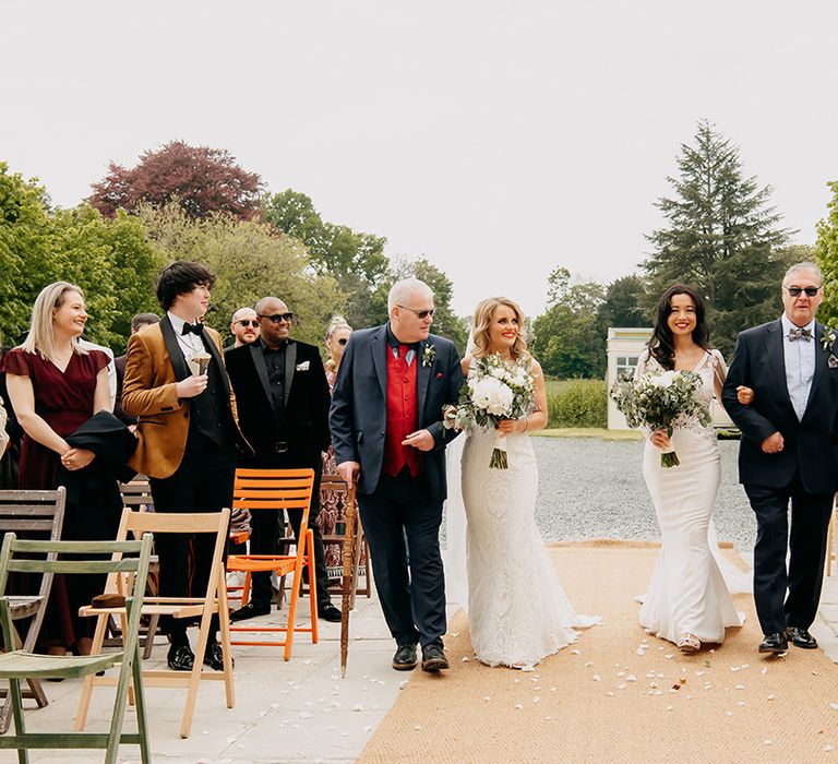 Father of the brides walk the brides down the aisle decorated with white flower petals at the Chateau De la Motte Husson