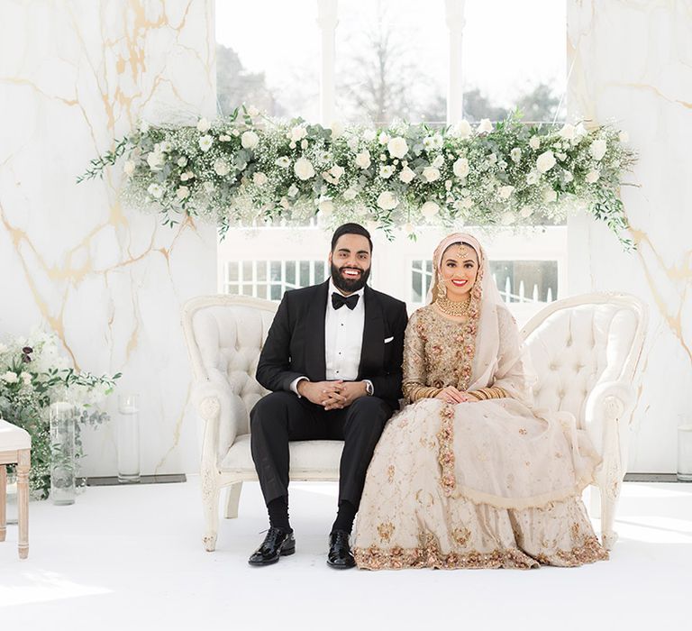 Bride wears traditional embellished saree on her wedding day as she sits beside groom wearing black tie in front of white florals 