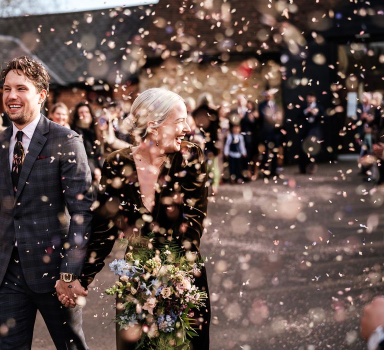 Groom holds brides hand as they walk through colourful confetti on their wedding day at Bradwell Abbey 