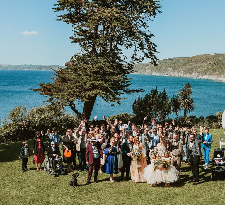 Bride & groom stand with their wedding party in front of the sea front in Cornwall for sunny wedding day outdoors