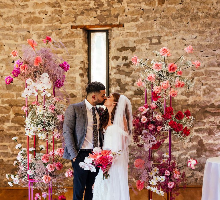 Bride & groom kiss between bright pink floral archway on their wedding day at Priston Mill