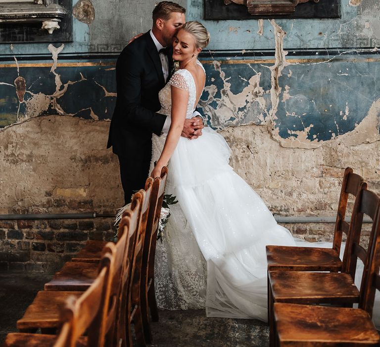 Groom kisses his brides cheek as festoon lighting hangs above them on their wedding day