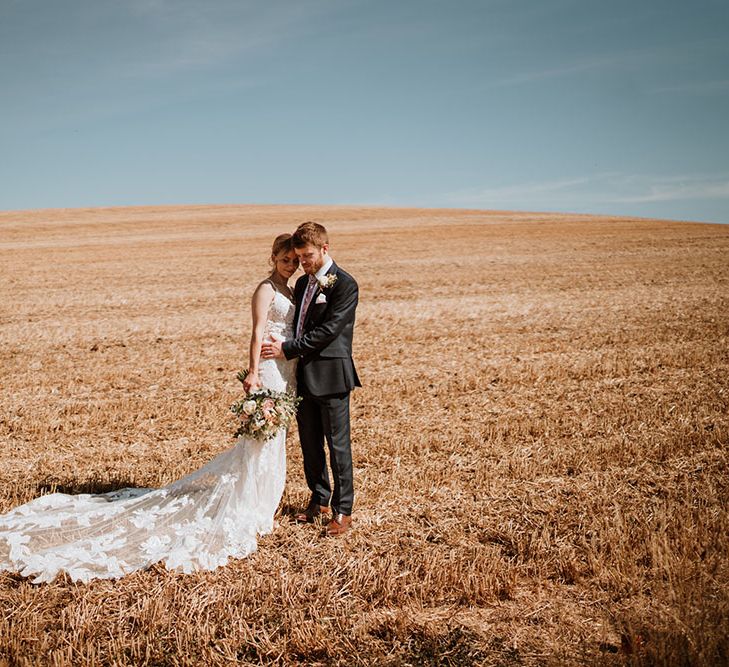 Bride wearing lace wedding dress & groom stand in golden field on the day of their wedding for couples portraits 