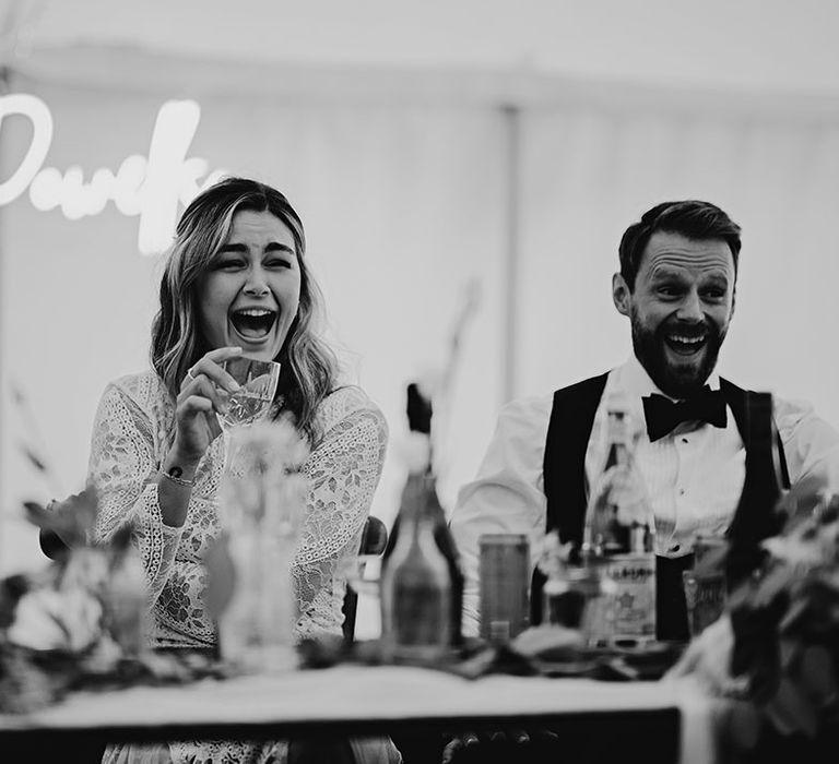 Bride and groom laugh during the wedding speeches as they sit in the marquee with a custom neon sign in the background 