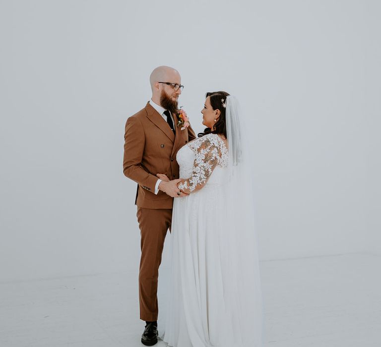 Bride wears lace embellished sheer sleeves and stands beside her groom in retro styled brown suit