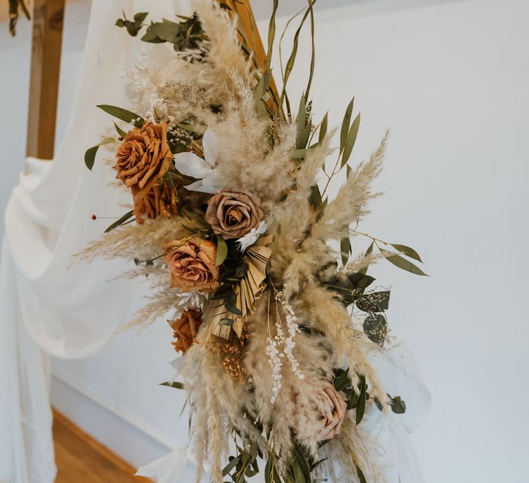 Wooden arch with large dried flowers, burnt orange and toffee coloured roses arrangement and white drapery hanging off