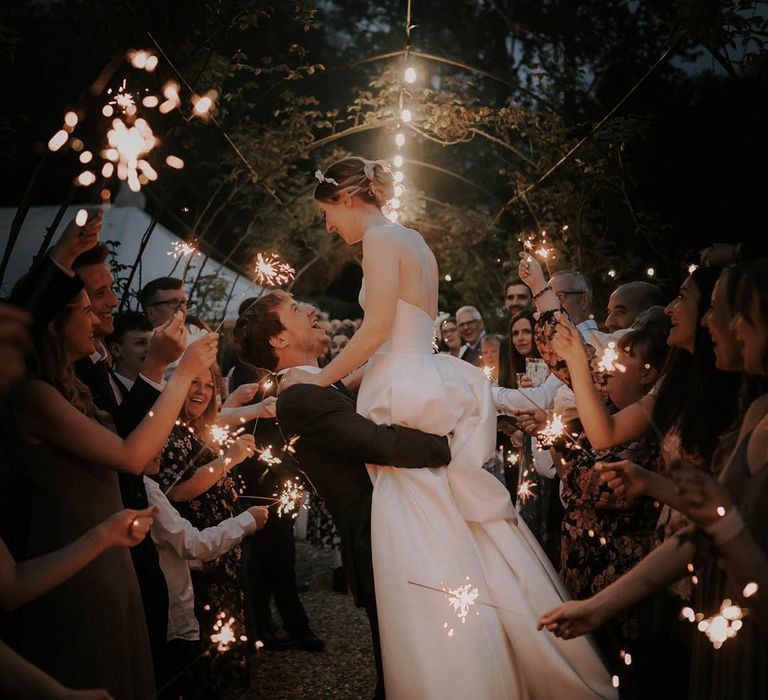 Groom in a dark grey suit lifts the bride for their sparkler send off 