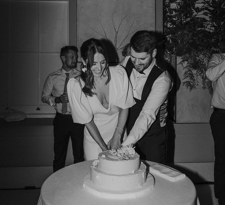 Bride in v neck wedding dress with puff sleeves and groom in white shirt and waistcoat cutting two tiered wedding cake with floral decorations 