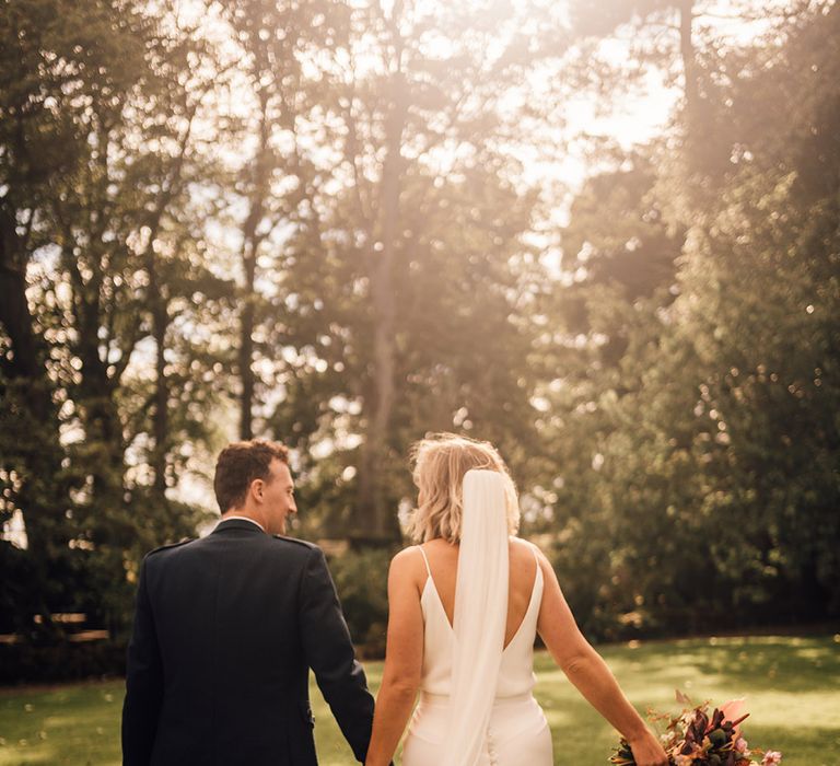 Bride in cathedral veil walks with her groom during golden hour outdoors in Scotland