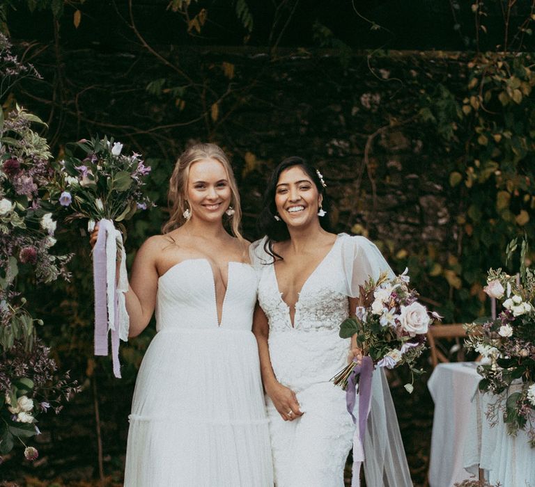 Brides in complimenting wedding dresses and Dr Martens boots smiling and laughing holding mixed floral bouquets with white carnations, purple tulips, roses and foliage 