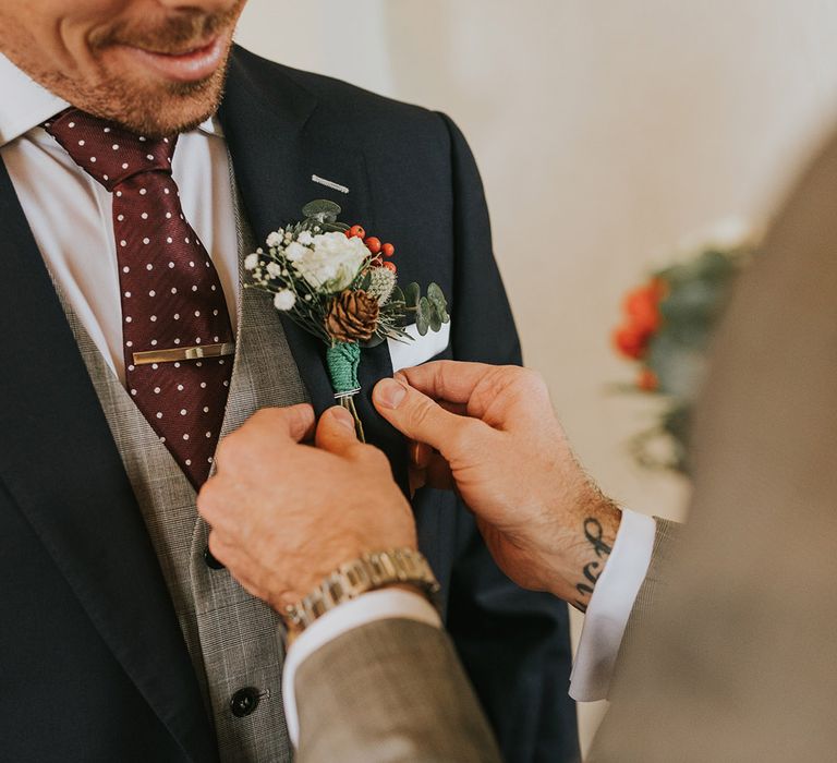 The groom in a grey suit helps the other groom by putting on his buttonhole made up of white flowers, red berries, and a pinecone for their winter wedding 