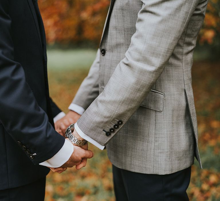 Groom in navy suit jacket holding hands with groom in grey suit jacket 