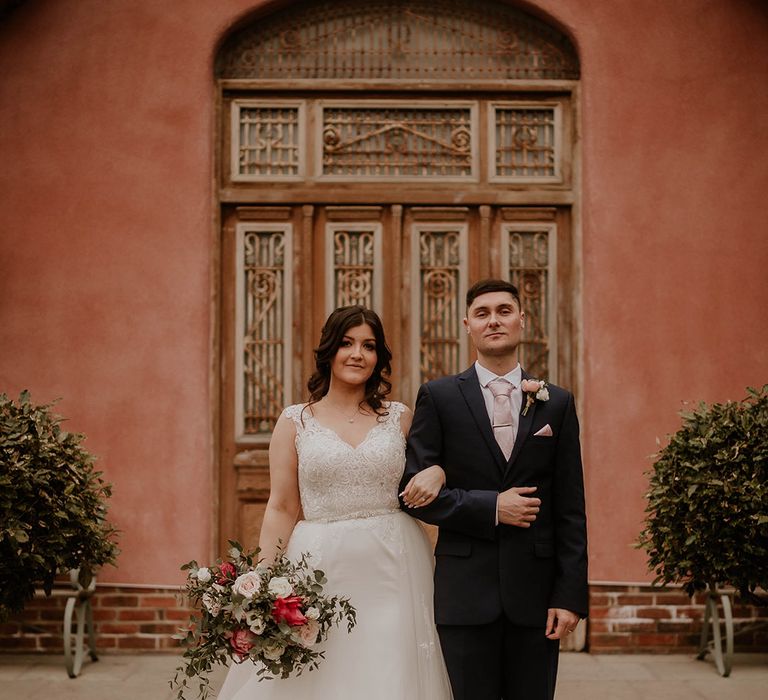 Bride in lace short sleeve wedding dress holds bridal bouquet and stands beside her groom in three piece suit 