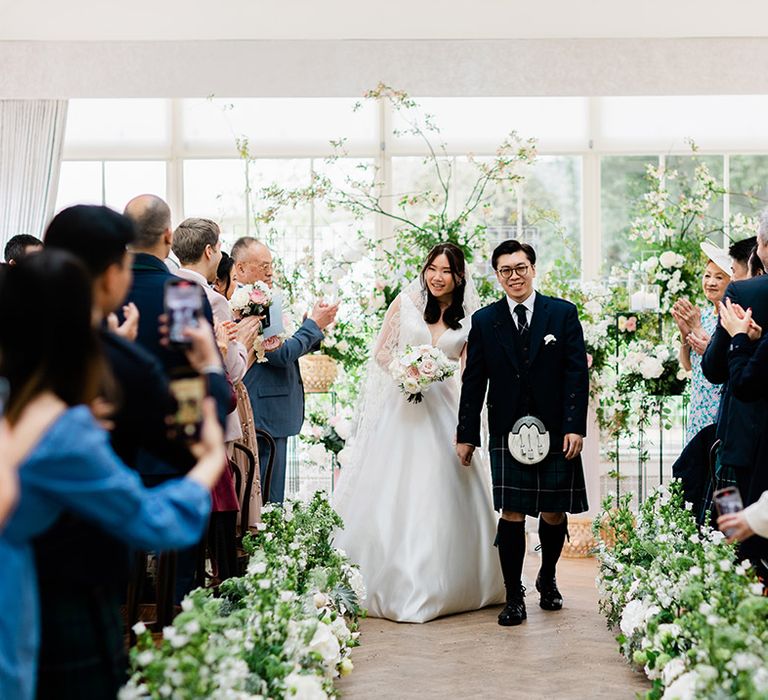 Bride in Princess wedding dress walks down the aisle with her groom in tartan green kilt