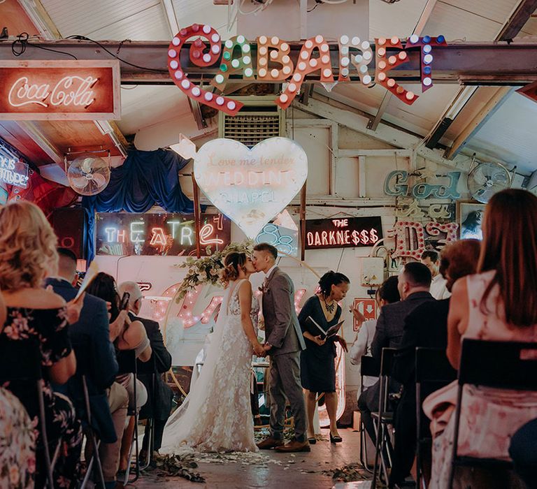 Groom in grey three piece wedding suit with yellow suit and yellow pocket square and bride in sleeveless plunge lace wedding dress  and cathedral length embroidered veil kissing at the alter of Gods Own Junkyard 