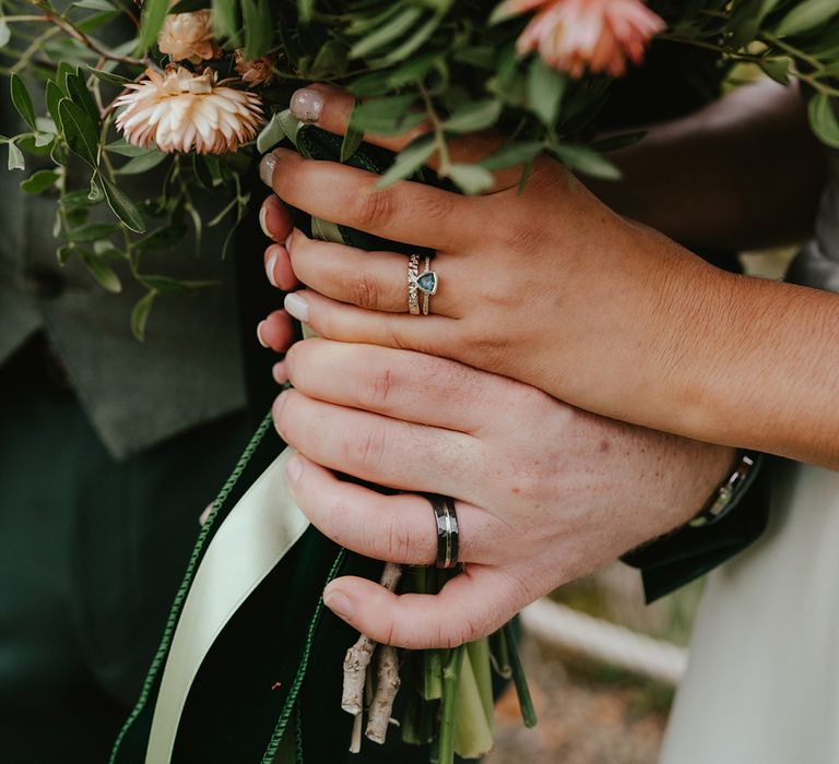 Bride & groom hold bridal bouquet showcasing their wedding bands and engagement ring