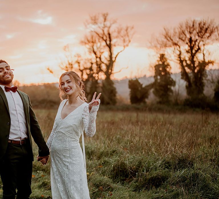 Bride in boho long sleeve lace wedding dress with tassels and groom in classic black suit with burgundy bowtie doing peace signs on the grounds of Kent barn wedding venue 