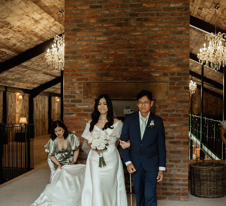 Bride in off the shoulder Eva Lendel wedding dress being walked down the aisle by the father of the bride in a blue suit with sage green tie 