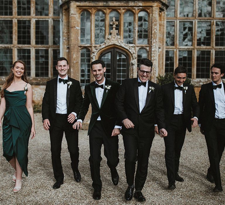 Groom and groomsmen in traditional black tuxedos with a groomswoman in a dark green cowl neck bridesmaid dress 