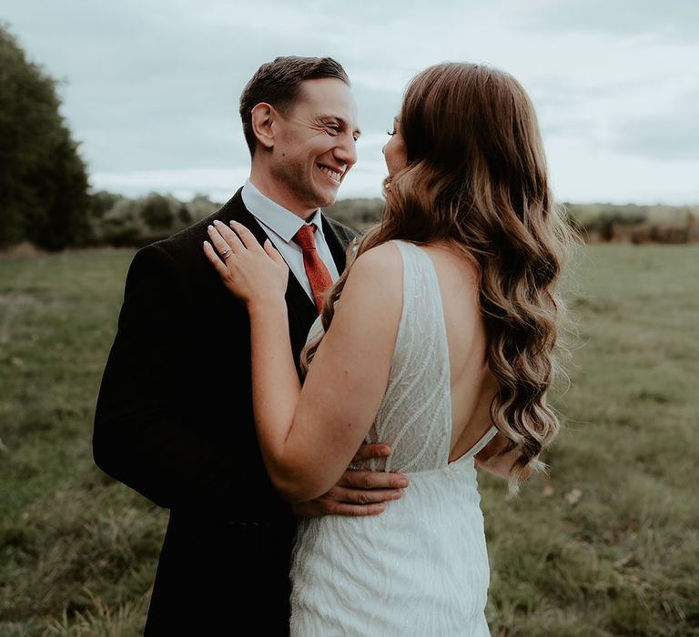 Groom smiles widely wearing a black suit and orange tie embracing the bride in a sequin wedding dress with curled hair 