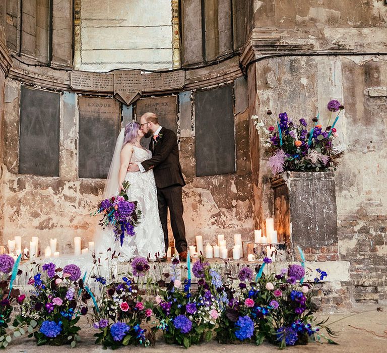 Groom in mocha brown textured suit with deep green tie and pink and purple boutonniere embracing and kissing bride in dainty leaf 3d appliqué sheer bridal gown and cathedral length veil at the alter of Asylum Chapel London surrounded by pillar candles, purple, blue, pink and white flower arrangements with dried flowers and bunny ears