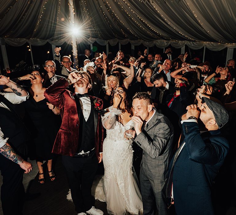 The wedding guests all take a shot together at the wedding marquee reception with fairy lighting 