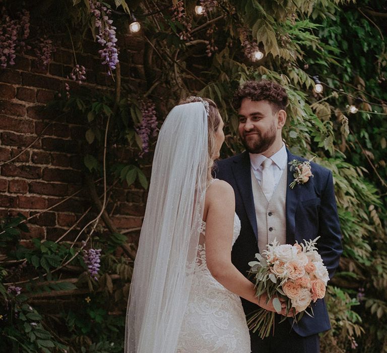 The bride in a fitted lace wedding dress wearing a cathedral length veil holding a pale pink and white rose bouquet facing the groom in a navy suit 