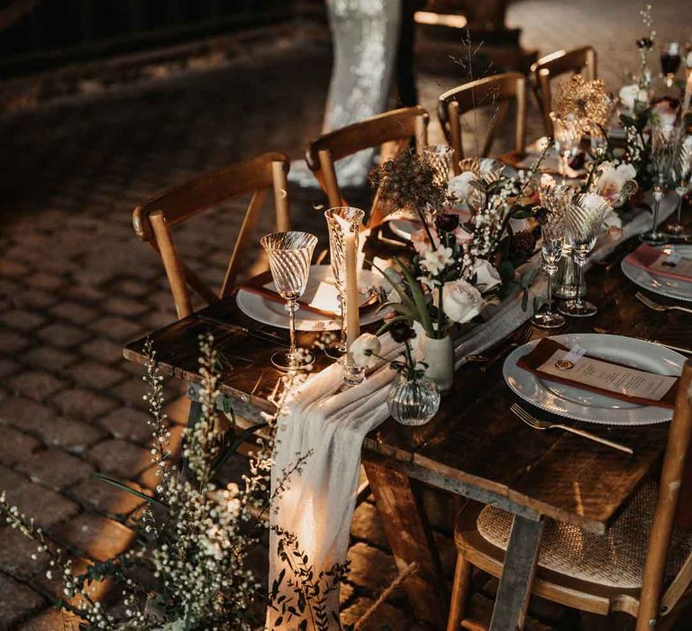 Bride and groom dancing by ustic luxe wedding tablescape with white fabric table runner, large foliage, baby's-breath, garden rose and eucalyptus floral arrangements, tapered candles and ceramic vases 