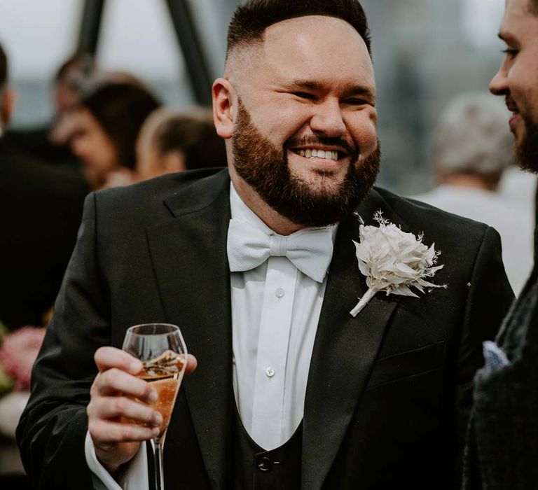 Groom wearing classic black three piece tuxedo with white bow tie and white garden rose, lavender twig and dried foliage boutonniere drinking champagne and laughing 