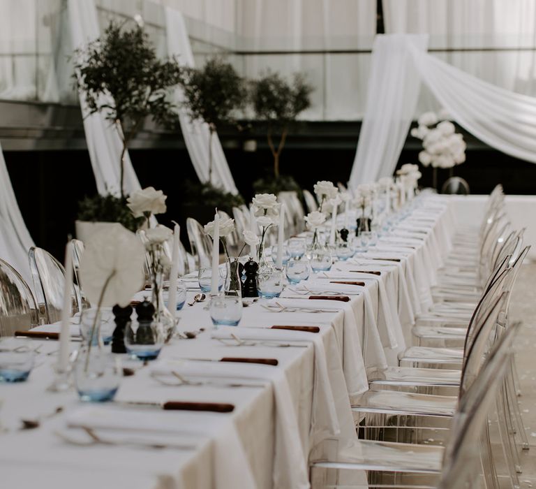 Acrylic wedding chairs with long banquet tables decorated with white tablecloths, white roses, white taper candles, and blue ombre glassware 