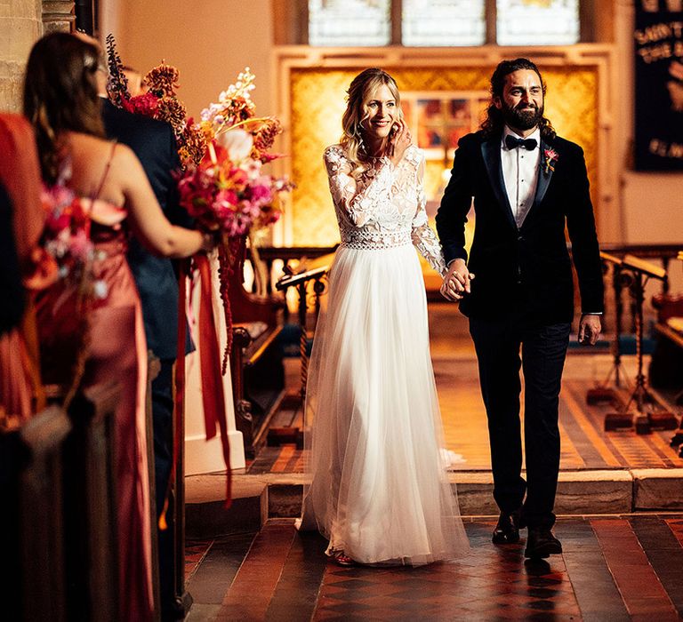 Bride in lace long sleeve wedding dress walking back down the aisle with groom in black tie suit 