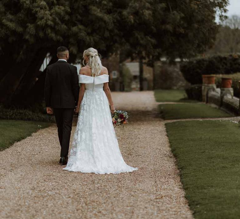 Bride in off the shoulder lace wedding dress holding colourful spring bridal bouquet walking through the grounds of Frensham Hall Manor House wedding venue with groom in classic three piece grooms tuxedo with colourful spring boutonniere 