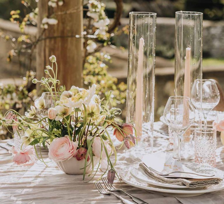 Blush pink tapered candles in glass candlestick covers on spring inspired wedding tablescape with white ruffled wedding table cloth, white peony, pink garden rose, foliage and blush florals wedding table centrepieces and simple crockery at Euridge Manor wedding