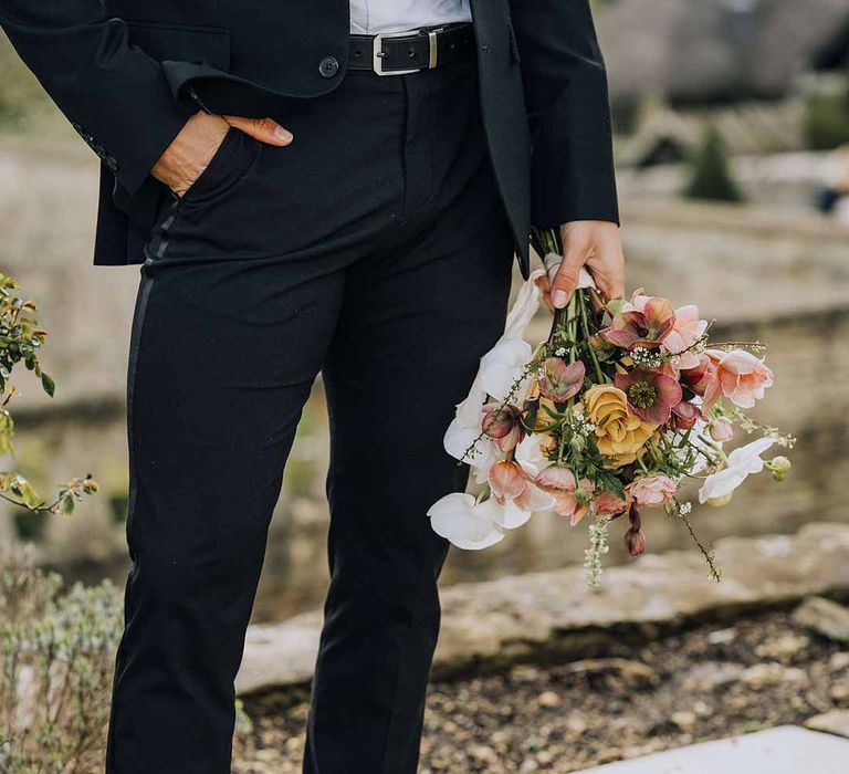 Groom holding blush toned wedding bouquet wearing classic black tuxedo and black dress shoes with red laces 