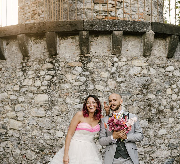 bride in a strapless layered wedding dress with pink edge and groom in a grey suit holding a pink orchid bouquet