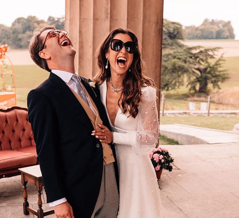 Groom in morning suit with yellow waistcoat and baby blue tie laughing with the bride in a pearl wedding dress and long sleeves wearing Gucci sunglasses