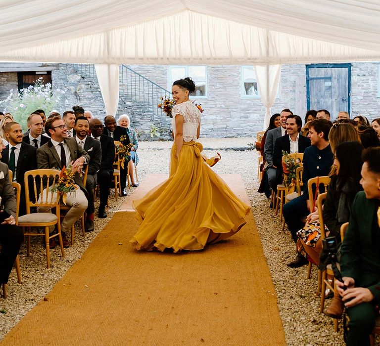 Bride in white mesh lace bridal crop top and yellow wedding skirt holding dried autumnal wedding bouquet with sunflowers, garden roses, pampas grass, eucalyptus, yellow tall kangaroo paws, orange osmanthus flowers, foliage and dried flowers walking down the aisle at Broadfield Court