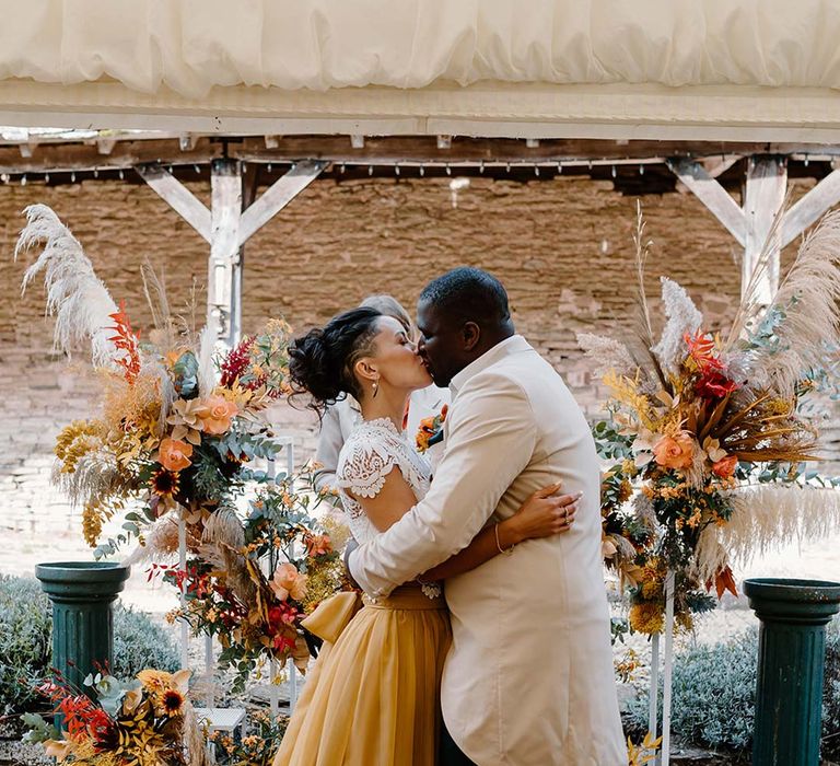 Groom in cream morning suit with forest green grooms waistcoat and trousers, forest green pocket square and orange boutonniere kissing bride in white mesh lace bridal crop top and yellow wedding skirt at the alter of Broadfield Court