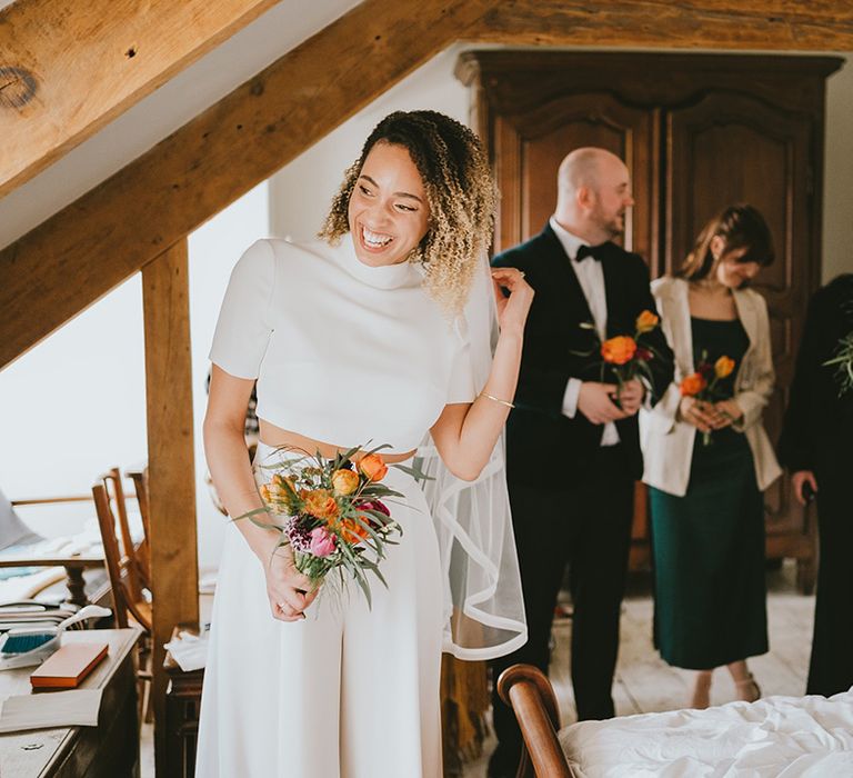 Bridal party in green bridesmaid dresses and green suit smiling with bride in short sleeve crop top and trousers bridal separates 