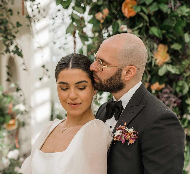 Bride in short sheer puff sleeve wedding dress with square neck and wrist loop with sheer pearl embellished bridal veil holding dried wildflower bouquet standing with groom in classic black tuxedo with bowtie and wildflower boutonniere at Buxted Park