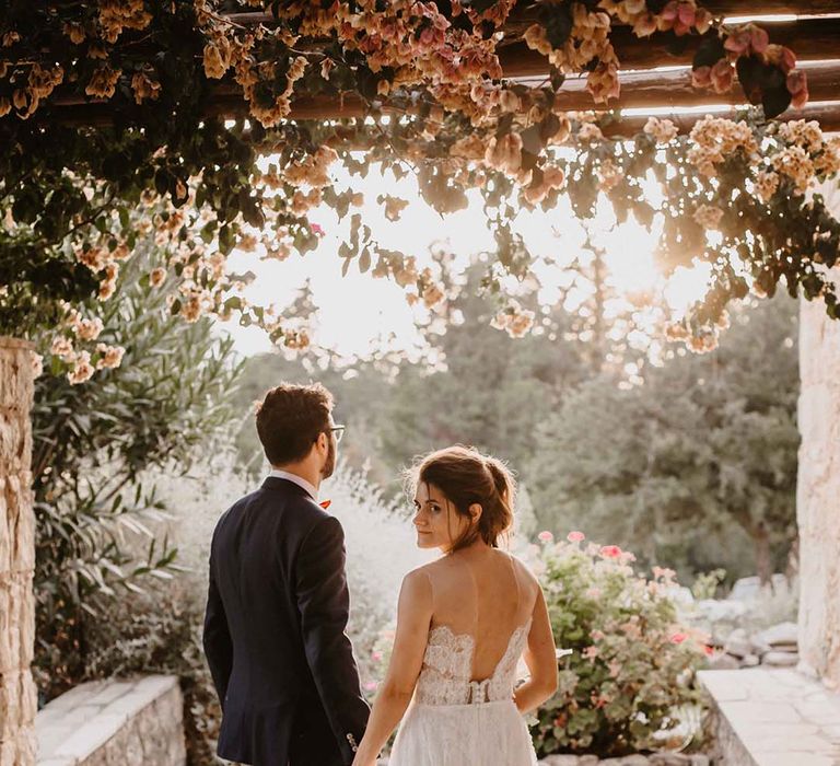 groom in grey chinos and navy blazer holding hands with his bride in a strapless lace wedding dress with low back design