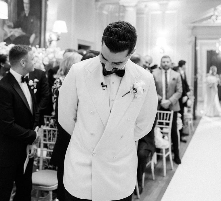 Groom in white double breasted suit jacket with black bow tie waiting for the bride at the altar 