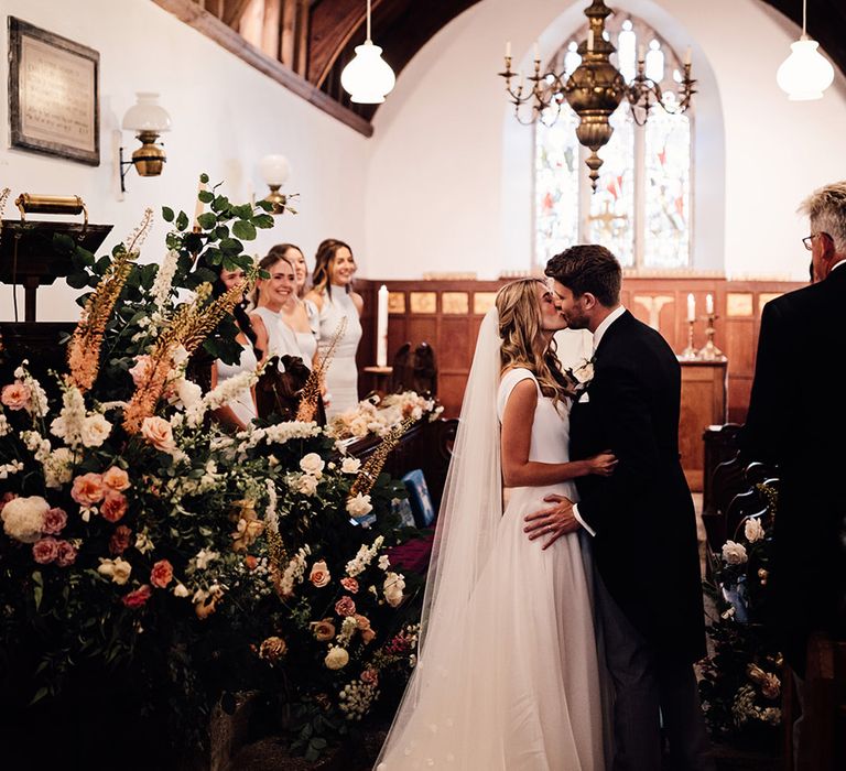 bride in a princess wedding dress and cathedral length veil and groom in a morning suit kissing at the church altar with large wedding ceremony floral arrangement 