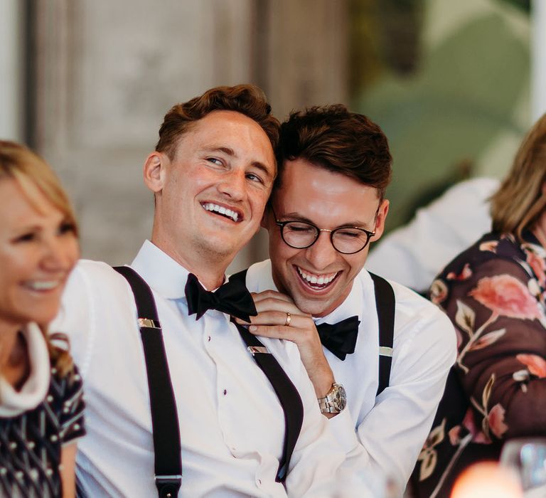 Two groom sit laughing at their wedding breakfast during the speeches 