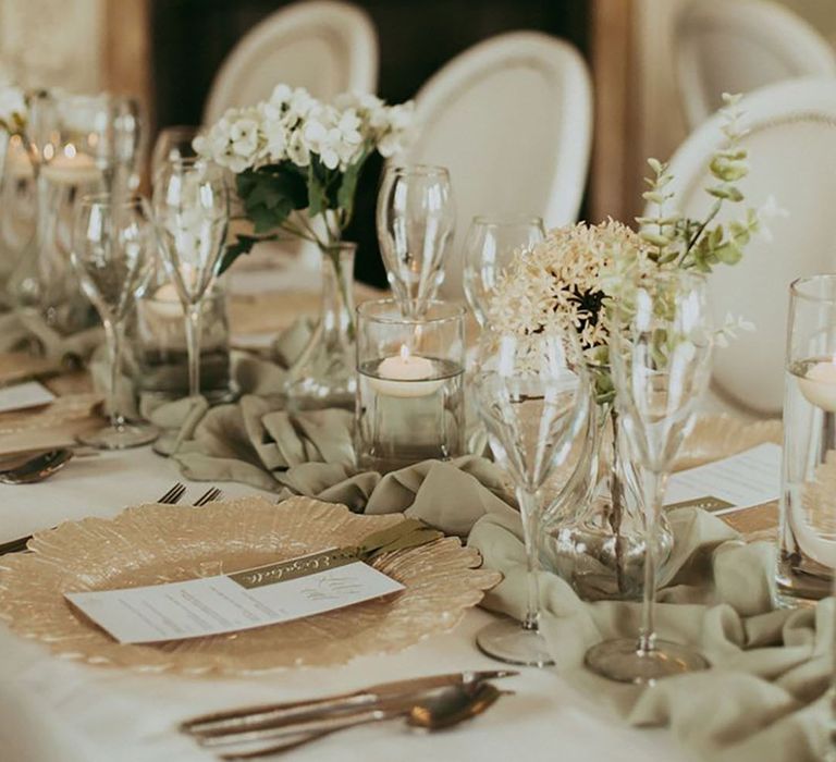 Gold flower wedding charger plate with sage green table runner with white wedding flowers and floating candles 